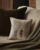A beige sofa with a dark brown cushion, a green knitted throw, and the Coral & Tusk Aspen Skiers Pillow—embroidered with winter-clad figures among bare trees—adds a charming touch of winter decor to the cozy setting.