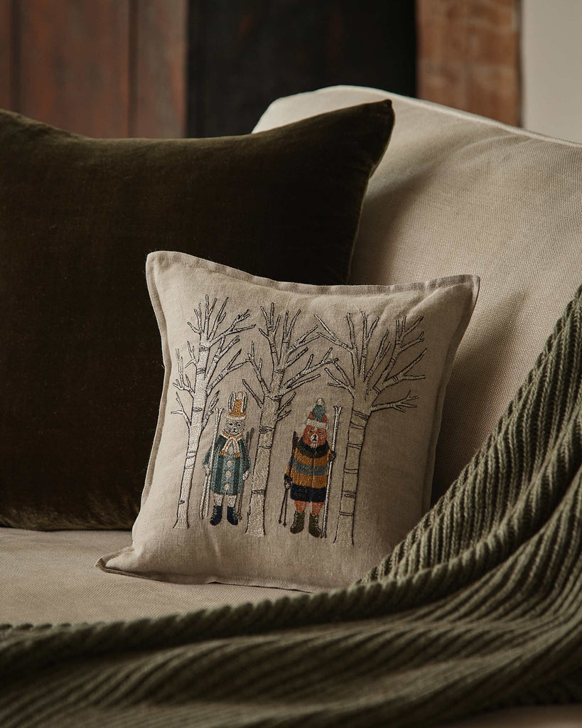 A beige sofa with a dark brown cushion, a green knitted throw, and the Coral & Tusk Aspen Skiers Pillow—embroidered with winter-clad figures among bare trees—adds a charming touch of winter decor to the cozy setting.