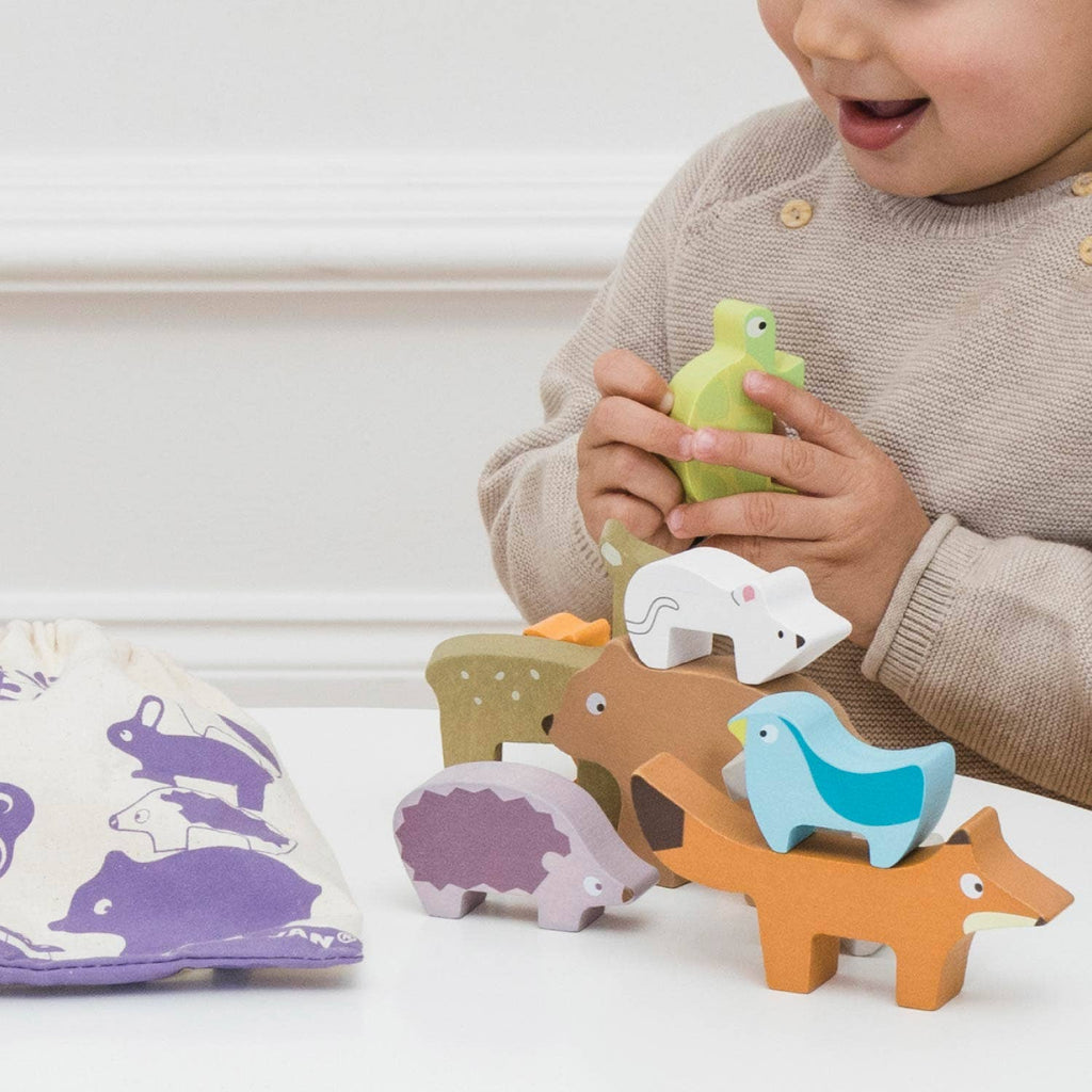 A young child plays with the Woodland Stacking Forest Animals & Bag, stacking colorful wooden animal pieces on a table. Nearby sits a cloth bag decorated with woodland animal prints. The child smiles and holds a green bird from the set. This sustainable toy encourages creativity and imaginative play.