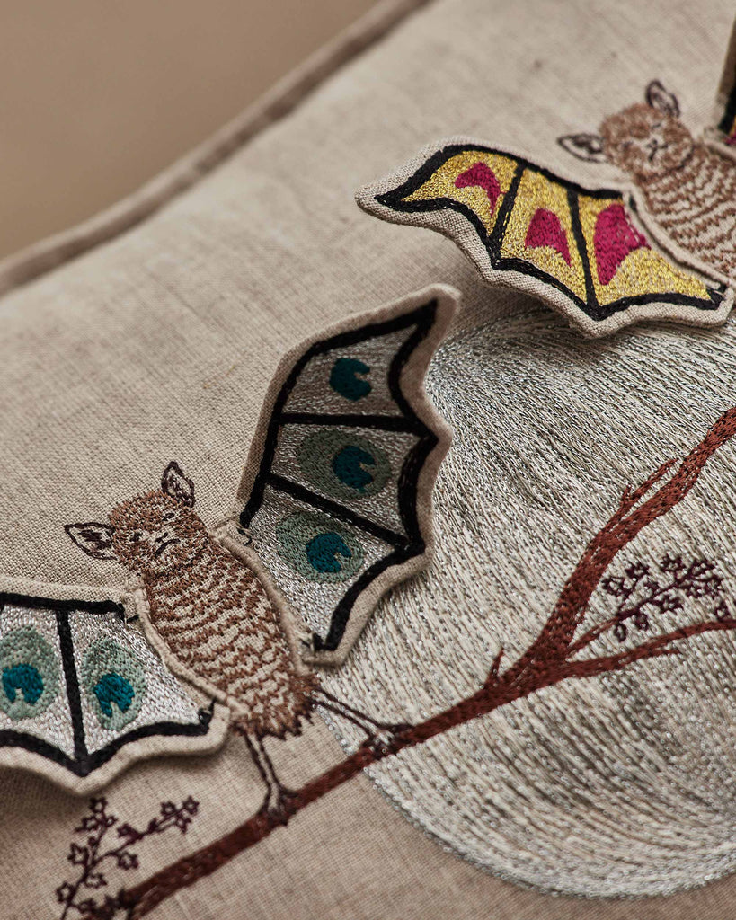 Close-up of the Coral & Tusk Bat Branch Pillow, featuring embroidered bats with butterfly wings on a brown branch against textured beige fabric and a moon-like design—perfect for a unique Halloween display. Ships in one week.