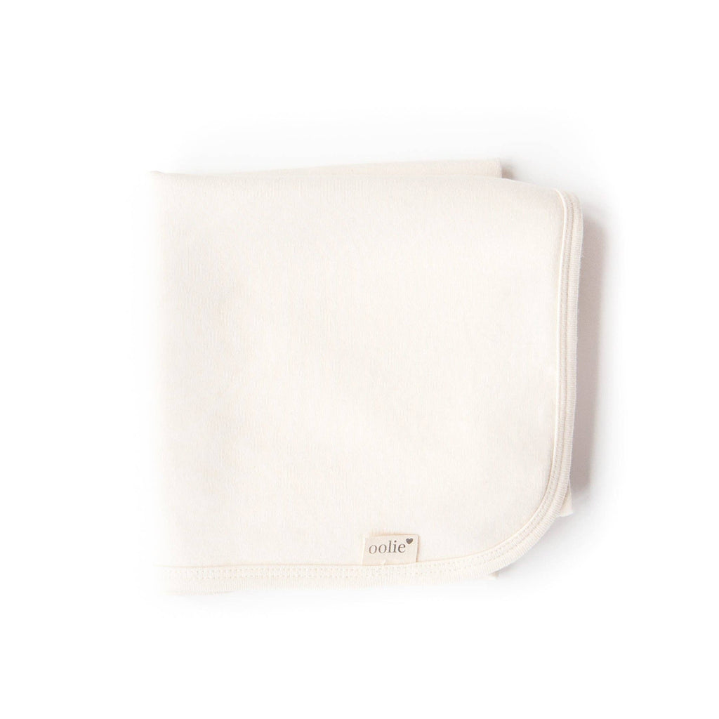 The Organic Baby Blanket – Natural is a soft, folded, off-white, chemical-free blanket with rounded corners and a small "oolie" fabric tag on the edge, shown on a white background.
