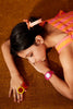 A girl in a pink-striped dress lies on a brown carpet, wearing the Mini Kyomo Watch - Ruby Papaya, colorful ring pops, and a peach ribbon in her dark hair. She looks relaxed and thoughtful.