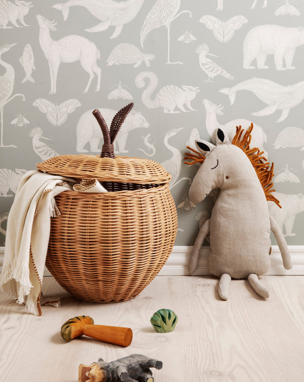 A cozy children's room corner featuring a Braided Apple Basket - Large with a lid and a fringe blanket, a stuffed dinosaur, and assorted plush toys on a wooden floor against a dinosaur-themed wallpaper background.