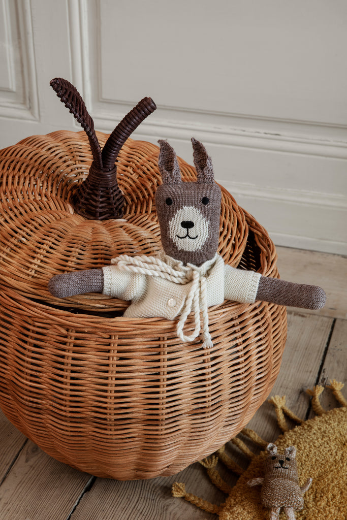 A knitted toy llama sitting inside a Braided Apple Basket - Large, with an upright lid featuring antlers. A smaller toy is on a rug next to the basket. The setting is cozy with a