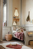 A cozy children's bedroom features a gray bed, playful face pillows, and a mauve quilt. Above, the Ferm Living Place Rack - Small displays decor and books. A wicker basket, soft rug, and cheerful wall art complete this warm and inviting space.