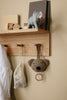 The Ferm Living Place Rack - Small, crafted from oak, organizes books, a white net bag, an elephant figurine, a hanging plush bear toy with ring, and a white shirt on pegs against a beige wall for stylish home storage.