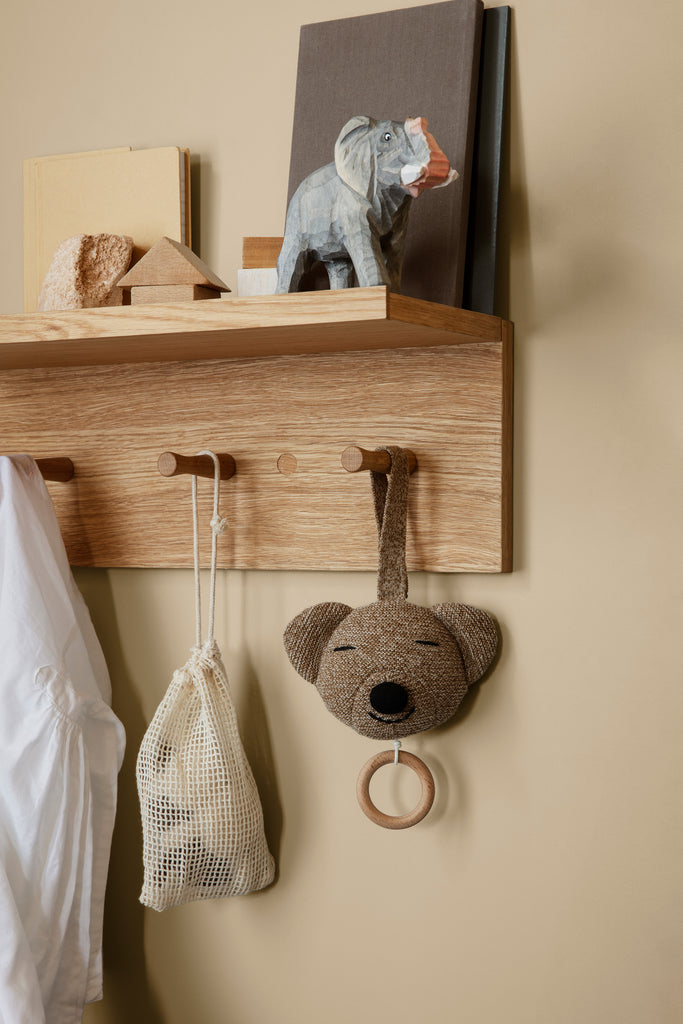 The Ferm Living Place Rack - Small, crafted from oak, organizes books, a white net bag, an elephant figurine, a hanging plush bear toy with ring, and a white shirt on pegs against a beige wall for stylish home storage.