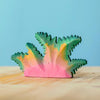 A small, colorful Bumbu Handmade Wooden Fern Bush, handcrafted and painted in shades of pink, yellow, and green, sits on a light wooden surface against a blue background.