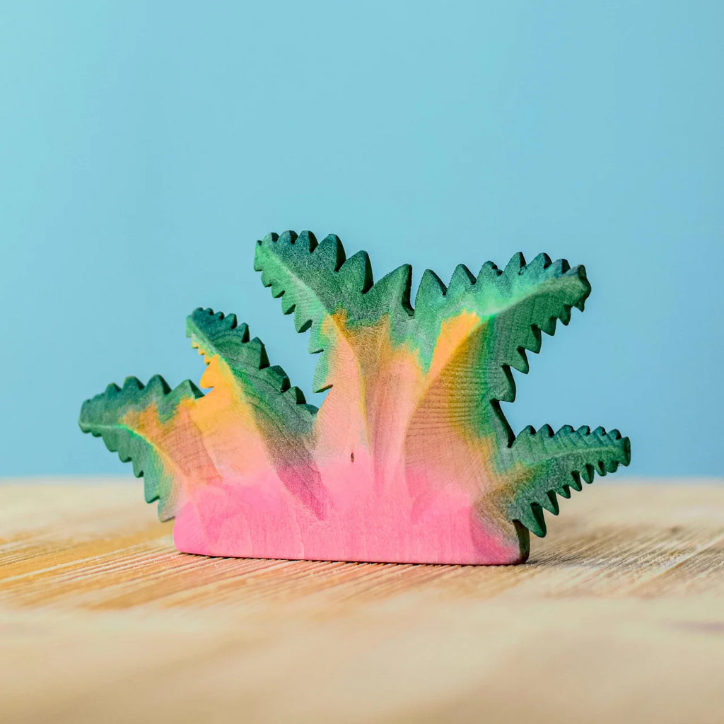A small, colorful Bumbu Handmade Wooden Fern Bush, handcrafted and painted in shades of pink, yellow, and green, sits on a light wooden surface against a blue background.