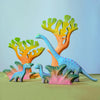 Three toy dinosaurs, painted blue with purple spots, stand among colorful toy trees and handcrafted toy plants, including the Bumbu Handmade Wooden Fern Bush, on a plain surface with a soft blue background.