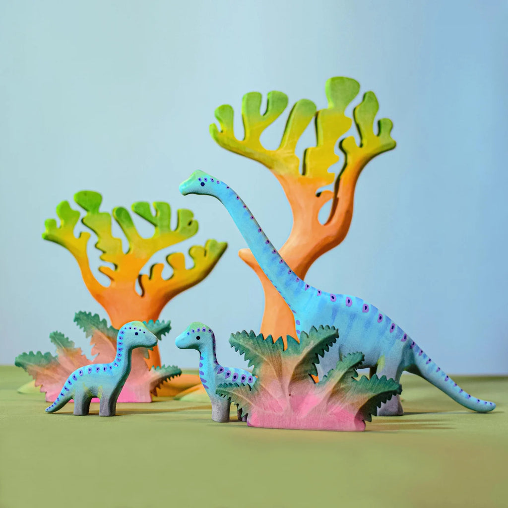 Three toy dinosaurs, painted blue with purple spots, stand among colorful toy trees and handcrafted toy plants, including the Bumbu Handmade Wooden Fern Bush, on a plain surface with a soft blue background.