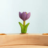 The Bumbu Handmade Wooden Festive Crocus—a small, carved purple tulip with green leaves—stands on a light wooden surface against a pale blue background, making a charming Montessori toy and eco-friendly decorative piece.