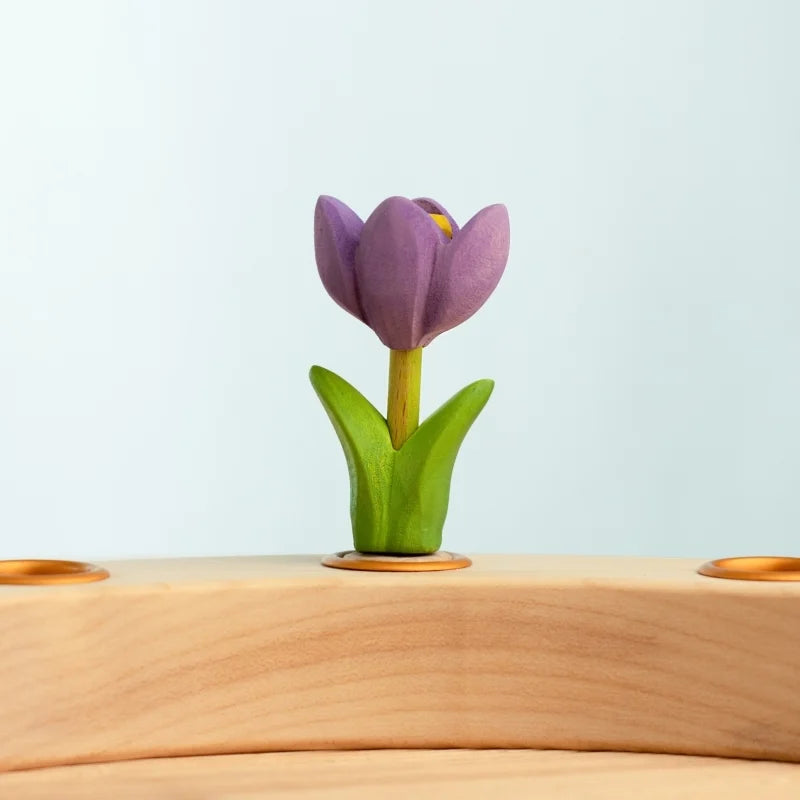 The Bumbu Handmade Wooden Festive Crocus—a small, carved purple tulip with green leaves—stands on a light wooden surface against a pale blue background, making a charming Montessori toy and eco-friendly decorative piece.