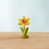 The Bumbu Handmade Wooden Festive Daffodil, featuring yellow petals, a red-orange center, and green leaves, stands upright on a wooden surface against a light blue background.