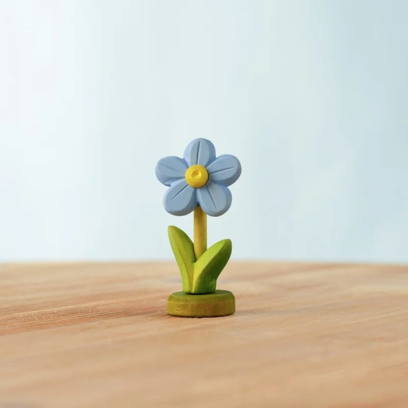 The Bumbu Handmade Wooden Festive Forget-Me-Not features a small blue clay flower with five petals, a yellow center, and green leaves, standing upright on a handcrafted wooden celebration ring against a softly blurred backdrop.