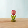 The Bumbu Handmade Wooden Festive Tulip, a small pink toy with green leaves, stands upright on wood against a blurred background—an eco-friendly, charming choice for Montessori-inspired play.