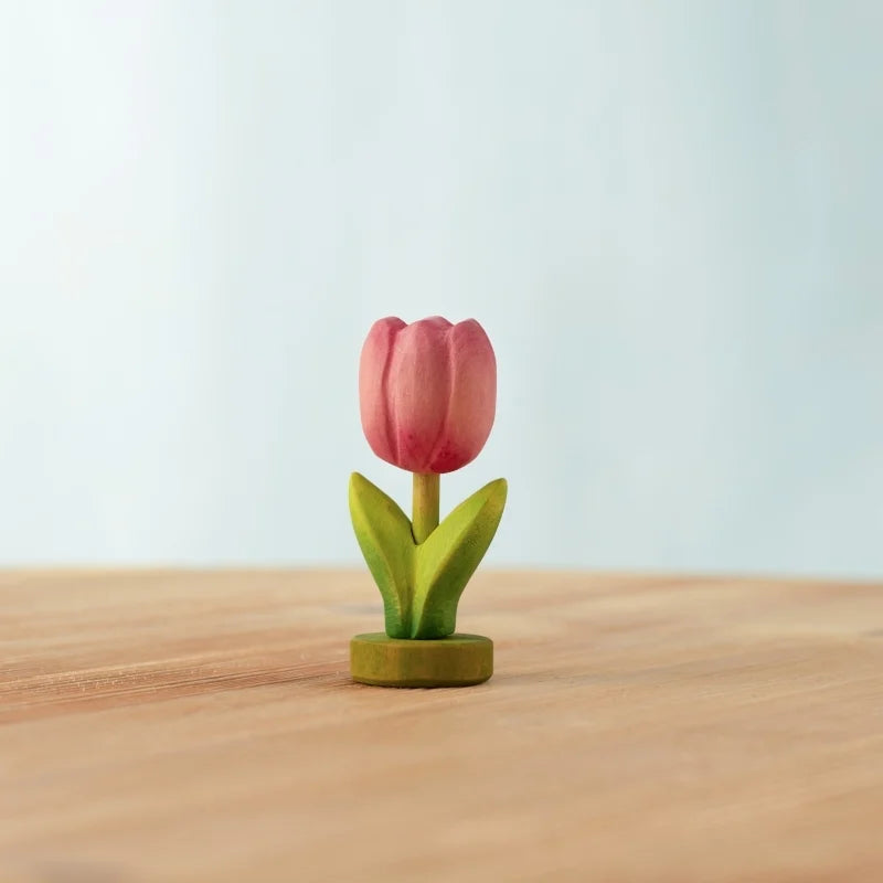 The Bumbu Handmade Wooden Festive Tulip, a small pink toy with green leaves, stands upright on wood against a blurred background—an eco-friendly, charming choice for Montessori-inspired play.