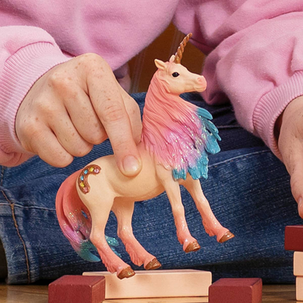 A person in pink sleeves holds a Schleich Marshmallow Mare Unicorn, featuring a pink and blue mane, standing the limited edition figure on wooden blocks.