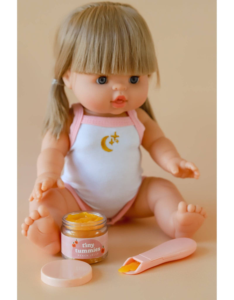 A doll with blonde hair and bangs wears a white and pink outfit featuring a moon and star design. In front of her is an open jar of Tiny Tummies Peach Jelly Food, accompanied by a small pink magic spoon from Tiny Harlow. The background is a soft beige.