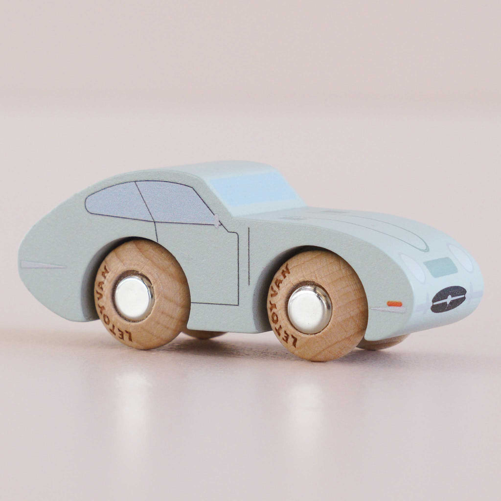 The No. 2 GT15 Jude | Made in England is a small, light blue wooden car with natural wood wheels, resting on a light pink surface. This charming vintage toy features simple painted details like windows and headlights, making it a delightful gift for kids.