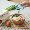 A hand pours water into a wooden bowl containing a Ramen Bowl Playset (ships in approximately one week) with plastic egg, narutomaki, and green lettuce. Fresh eggs, a leek, and wooden chopsticks are nearby on a checkered cloth. An egg carton is also in the scene, all crafted from natural non-toxic materials for sensory play food fun.