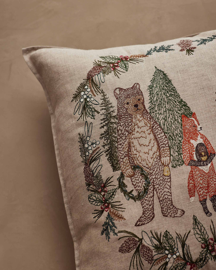 A Coral & Tusk Christmas Cheer Wreath Pillow featuring a linen cover embroidered with a bear and a fox standing within a holiday wreath of pine branches, berries, and mistletoe.