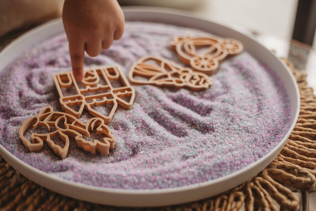 A child’s hand points at a wooden castle shape on purple sensory sand, with additional wooden shapes of a unicorn, crown, and dress also visible on the tray—all part of the Princess Cutter Set - Eco Playdough Tool. This enchanting and eco-friendly playdough cutter collection is designed to inspire imaginative princess-themed play while caring for the planet.