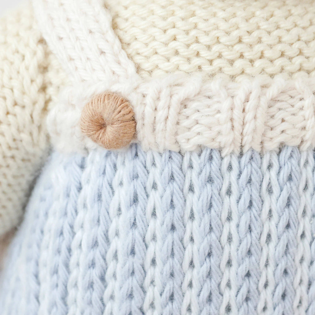 Close-up of Cuddle + Kind Benjamin the Lamb’s hand-knit outfit: a cream 100% cotton sweater with brown button paired with light blue textured overalls. Each detail is lovingly handmade by artisans in Peru.