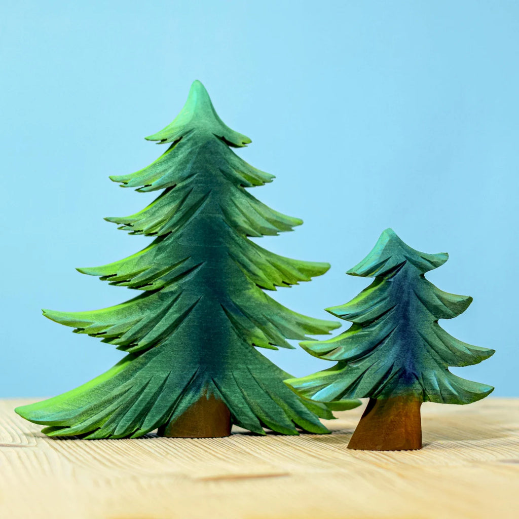 The Bumbu Handmade Wooden Fir Tree Set, featuring one tall and one short handcrafted wooden tree with green painted branches, stands on a light wooden surface against a light blue background.