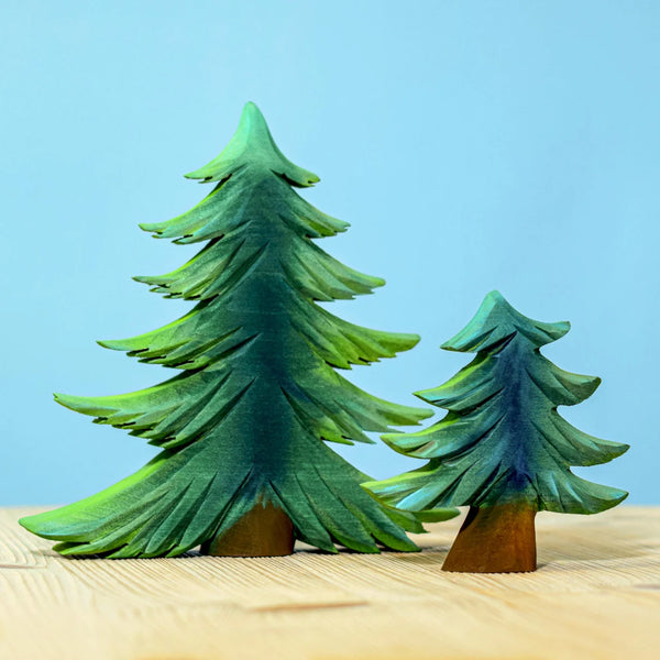 The Bumbu Handmade Wooden Fir Tree Set, featuring one tall and one short handcrafted wooden tree with green painted branches, stands on a light wooden surface against a light blue background.