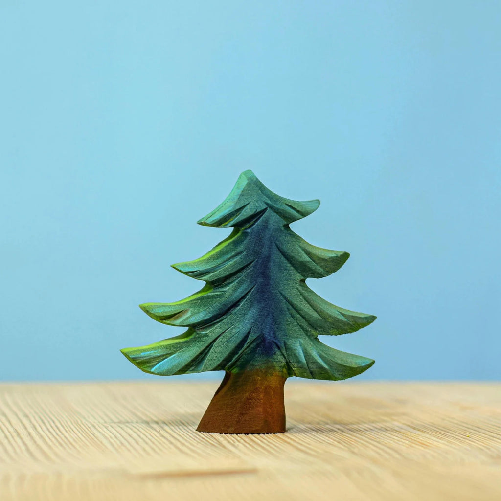 A small, handcrafted piece from the Bumbu Handmade Wooden Fir Tree Set—painted green and brown—stands on a light wooden surface against a plain blue background, making an eco-friendly addition to any nature-inspired toy collection.