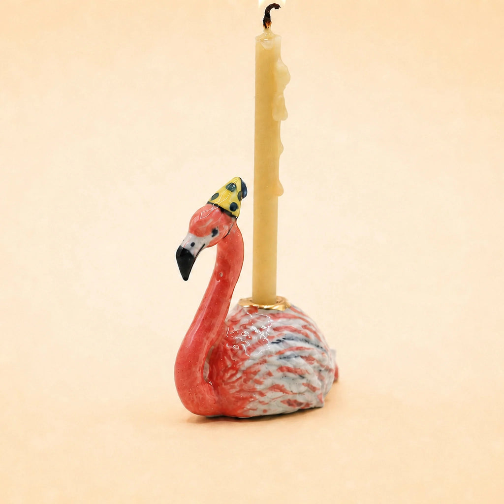 The Flamingo Cake Topper - Final Sale (ships in one week) features a ceramic flamingo with a yellow polka-dot hat and candle, making it an ideal handcrafted collectible or birthday gift.