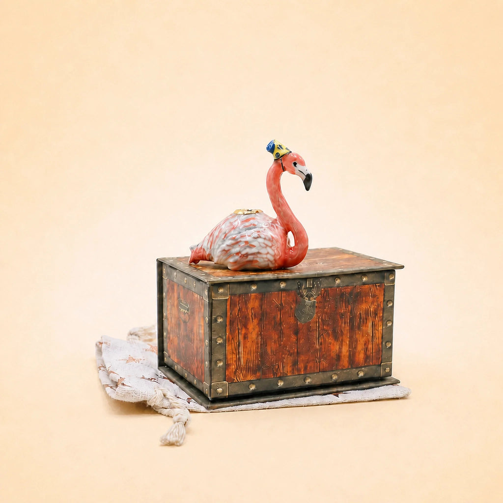The Flamingo Cake Topper - Final Sale (ships in one week) features a ceramic flamingo with a blue and yellow hat on a wooden chest—an adorable handcrafted keepsake on textured cloth.