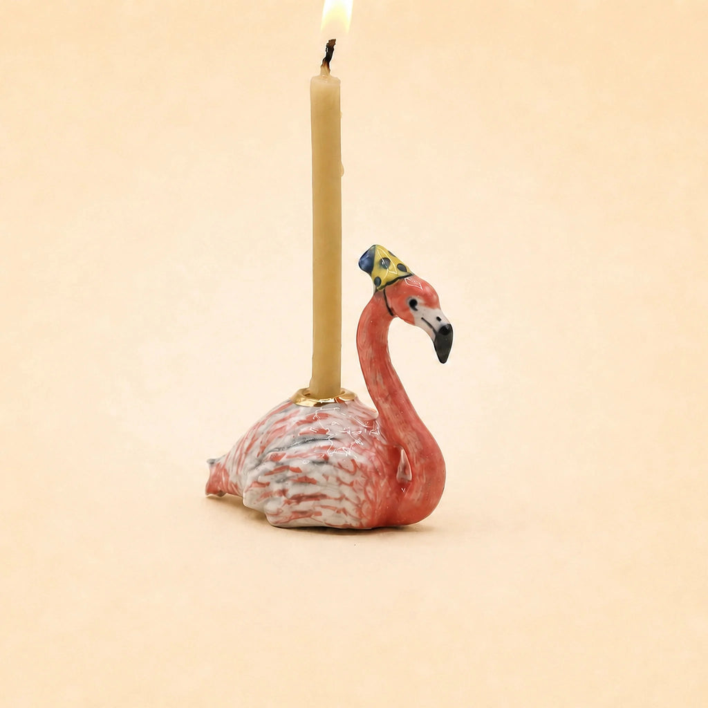 The Flamingo Cake Topper - Final Sale (ships in one week) is a porcelain figurine of a small flamingo in a yellow hat, carrying an upright candle on its back—a delightful handcrafted keepsake.