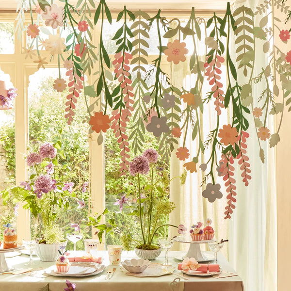 A sunny room with a table set for tea, surrounded by lush hanging plants and Meri Meri Floral Paper Backdrop, with a view of a garden through a window.