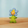The Bumbu Handmade Wooden Flower Child Forget-Me-Not, a small wooden figurine with a green body, smiling face, and blue five-petal flower with yellow center around its head, stands on a light wooden surface against a pale blue background.