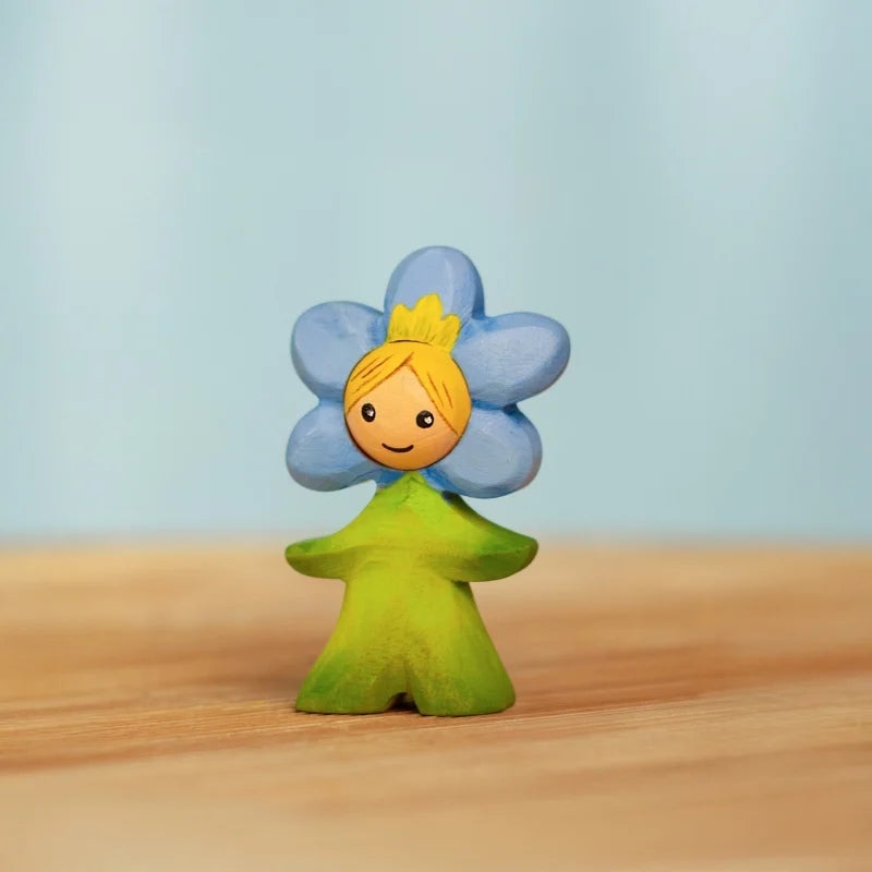 The Bumbu Handmade Wooden Flower Child Forget-Me-Not, a small wooden figurine with a green body, smiling face, and blue five-petal flower with yellow center around its head, stands on a light wooden surface against a pale blue background.