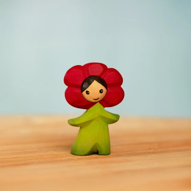 The Bumbu Handmade Wooden Flower Child Poppy is a cheerful figurine with a red flower-shaped head and green body, ideal for small world play, inspired by Waldorf toys—perfect on a light wooden surface against a blue background.