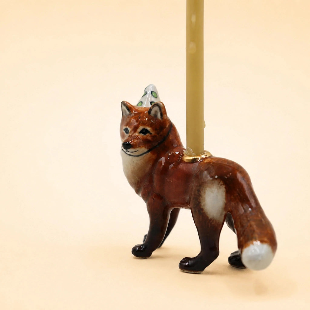A handcrafted “Fox Cake Topper - Final Sale” collectible ceramic figurine featuring a candle holder with a beige candle on its back, displayed against a plain light beige background.