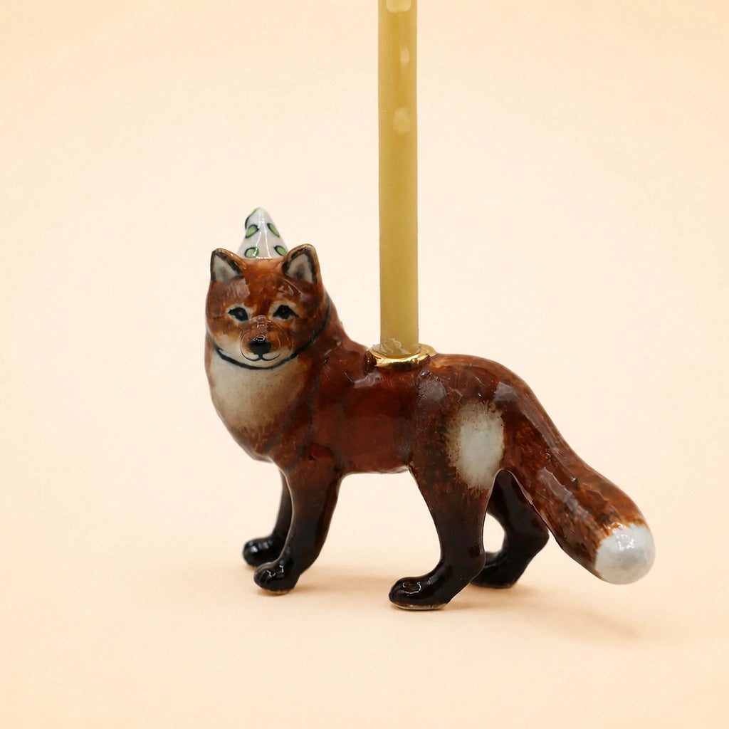 A charming handcrafted collectible, the Fox Cake Topper features a ceramic fox figurine sporting a festive party hat and balancing a slender upright candle on its back, set against a simple beige background.