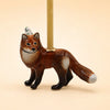 A handcrafted collectible, the Fox Cake Topper features a ceramic fox wearing a tiny party hat and holding a candle on its back, displayed against a beige background.