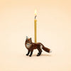 A lit beige candle sits in a holder shaped like a standing brown and white fox, set against a plain light beige background—this whimsical, handcrafted Fox Cake Topper (Final Sale) makes a charming collectible for any keepsake treasure chest.
