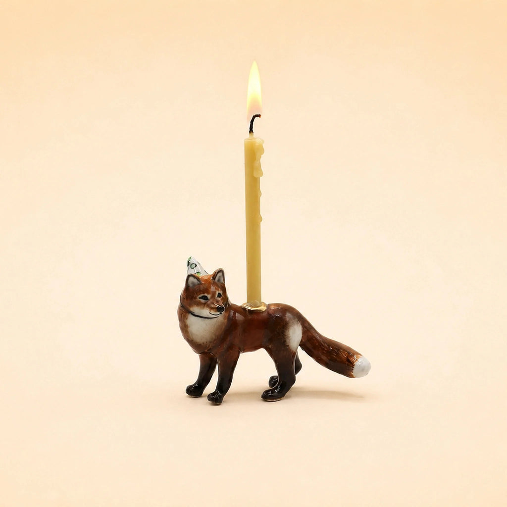 A lit beige candle sits in a holder shaped like a standing brown and white fox, set against a plain light beige background—this whimsical, handcrafted Fox Cake Topper (Final Sale) makes a charming collectible for any keepsake treasure chest.