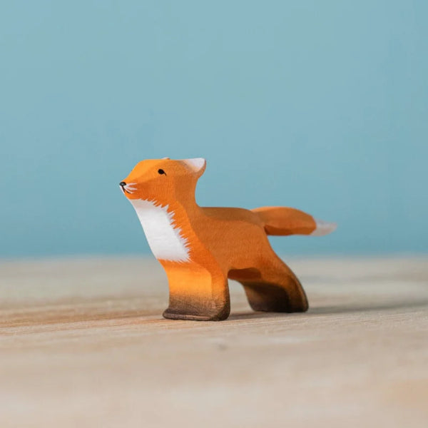 The Bumbu Handmade Wooden Fox Cub Curious, featuring orange, white, and brown hand-painted details, stands on a light wooden surface against a blue background for a charming eco-friendly toy.
