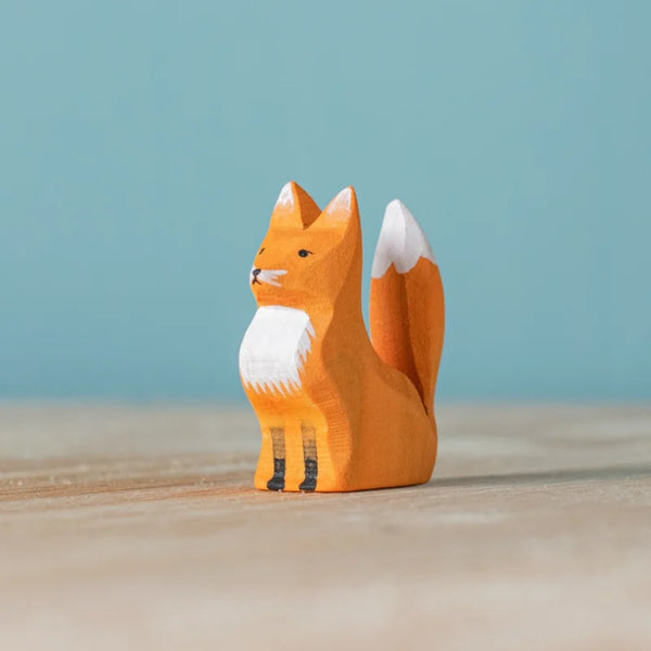 The Bumbu Handmade Wooden Fox Cub Sitting is a small, eco-friendly, hand-painted figurine with an orange body, white chest, and tail tip—perfect as a charming nature-inspired wooden toy or decor against a light blue background.