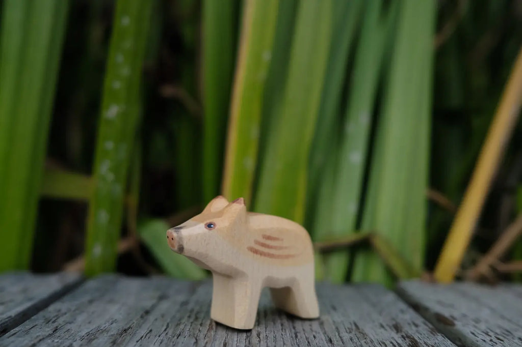 A small, hand-carved Handmade Holzwald Small Wild Boar - Head Up standing on a wooden surface against a backdrop of green leaves, perfect for imaginative play.