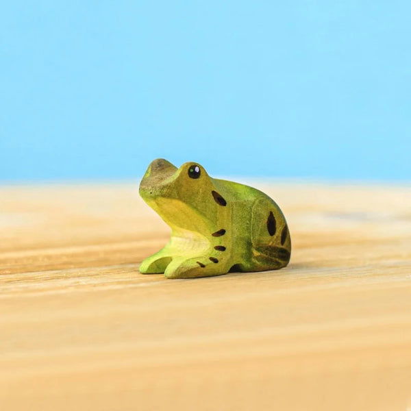 The Bumbu Handmade Wooden Frog, a small green handcrafted toy with dark spots, rests on a light wooden surface against a blue background.