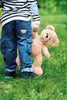 A toddler from behind holding a Fynn Teddy Bear in a green grassy area. The child wears a striped top and denim pants with a teddy bear design on the pocket.