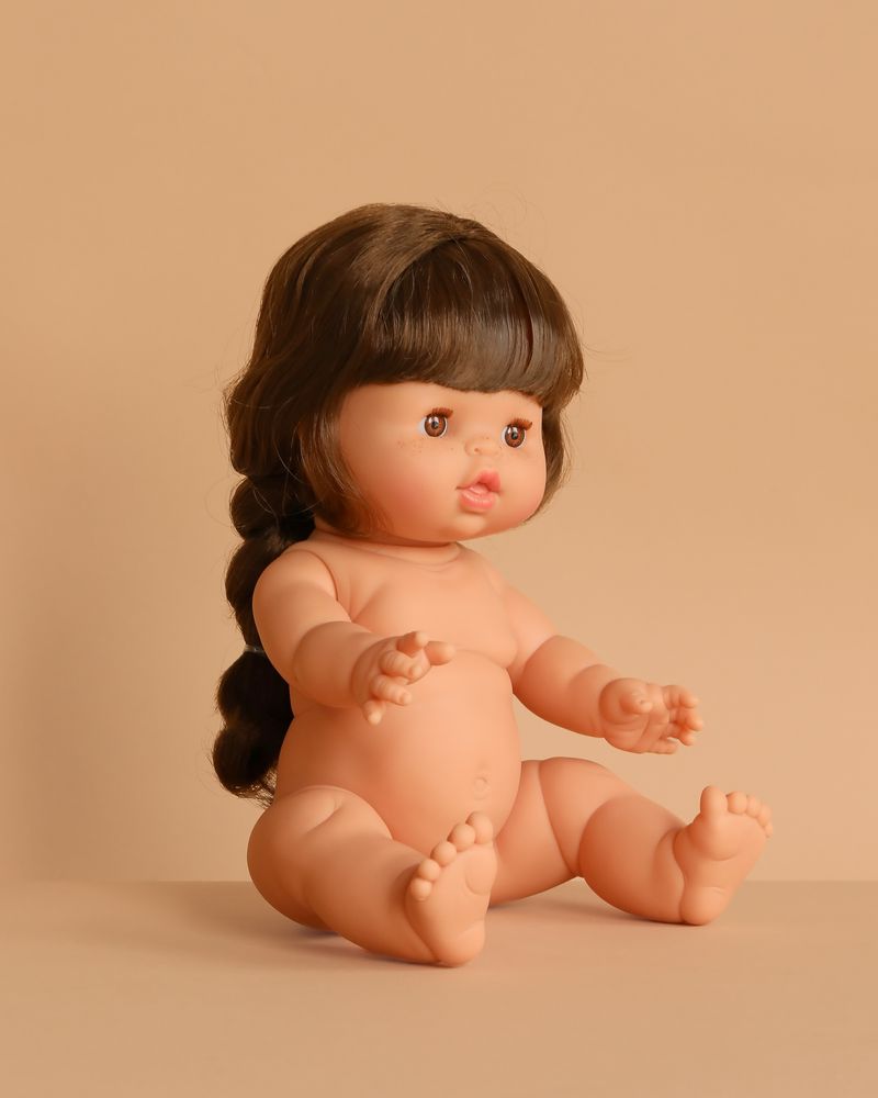 A Minikane Doll (13") - Valentine, featuring long brown hair in a braid, sits upright with extended arms and legs against a plain beige background. Clothing sold separately.