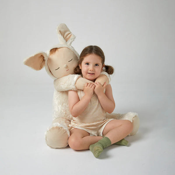 A young girl sits on the floor smiling, hugging the Giant Cozy Bunny Moppet—a plush bunny with floppy ears. The plain, light-colored background enhances this snuggly toy's charm and inviting appeal.